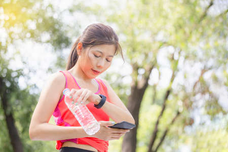 Young beautiful woman with plastic water bottle looking smartwatch and smartphone, Sport woman at...