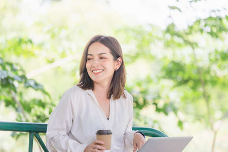 Young freelancer sitting with a laptop in a green park. Smiling modern woman sitting on bench while drinking takeaway coffeeの写真素材