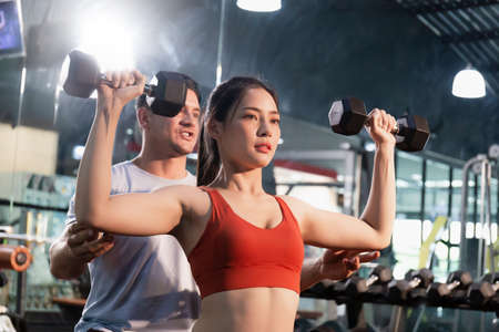 Young woman with dumbbells and personal trainer flexing muscles in gym, Sport and fitness conceptsの写真素材