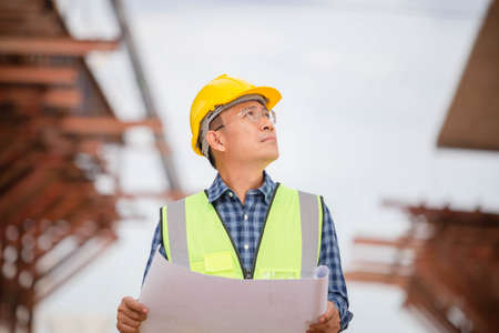 Asian construction engineer checking project at the building site, Architect with a blueprints at a construction site, Foreman worker in hardhat at the infrastructure construction siteの写真素材