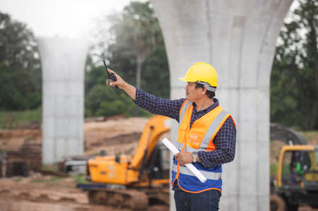 Construction engineer checking project at the building site, Architect with a blueprints at a construction site, Foreman worker in hardhat with Two-Way radio at the infrastructure construction siteの写真素材