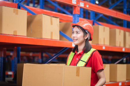 Female worker in hardhat holding cardboard box walking through in retail warehouse, Warehouse worker working in factory warehouseの写真素材