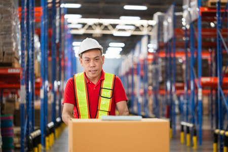 Cheerful warehouse worker checking inventory and working in warehouse, Worker man using hand pallet jack unloading package boxes at factory warehouseの写真素材