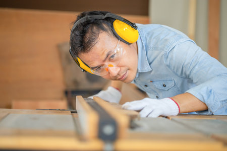 Woodworker man wearing safety gear using circular saw to cut a wood on workbench, Carpenter working...