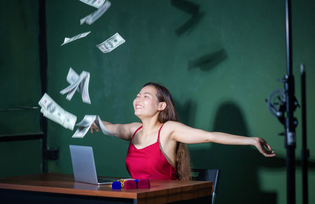Excited happy young woman sitting at table with dollars money flying overhead while raising handsの写真素材