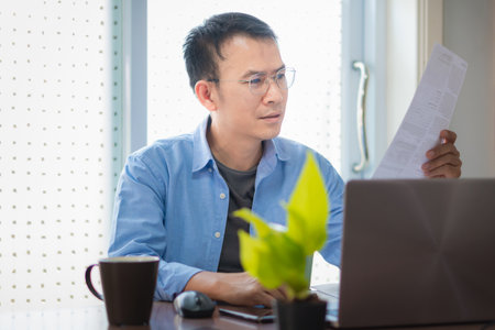 Asian man sitting at table drink coffee working with documents and laptop in homeの写真素材