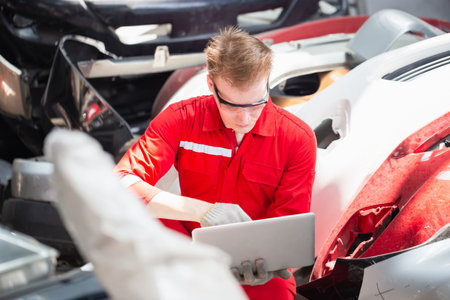 Mechanical man inspecting car parts stock on laptop computer while working in garage warehouse, Young mechanic with laptop in auto repair shopの写真素材