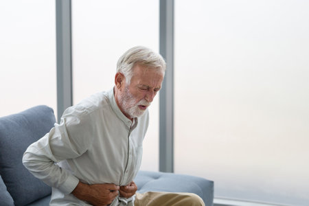 Unhappy senior man with stomach ache, man presses his hand to stomach from unbearable pain, mature man with stomach pain feeling unwell sitting in living roomの写真素材