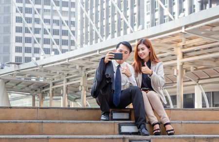 A joyful businessman and woman taking a selfie while sitting together on the stairs, Young Asian business people team discussing in the morningの写真素材