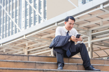 Asian businessman sitting on stairs and checking work on mobile smartphone outside office blurry city backgroundの写真素材