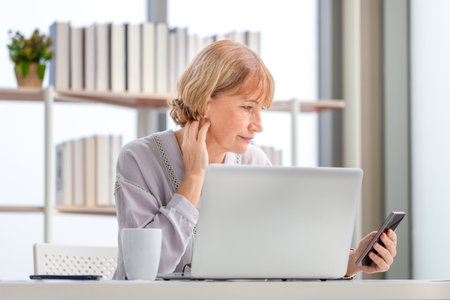 Portrait of woman using smartphone and laptop, Mature woman in living room with mobile phone browsing internet on modern computer gadgetの写真素材