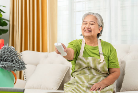 Smiling Asian senior woman with air conditioner remote control in living room, Woman switching on air conditioningの写真素材