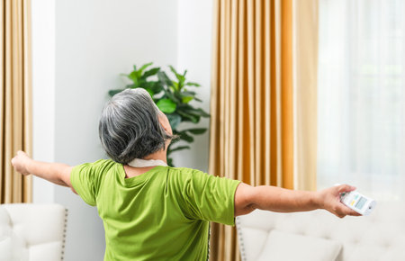 Back view of a relaxing senior woman standing with arms outstretched in living room, Woman switching on air conditioningの写真素材