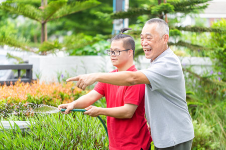 Cheerful senior man and grandson enjoying together are watering the plants with hose, grandfather with grandson working in the front yard gardenの写真素材