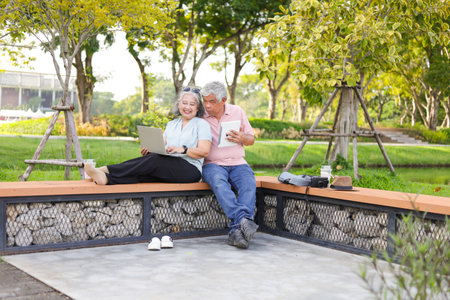 Cheerful senior couple sitting on a bench in park, Asian senior man and woman use laptop and digital tablet in parkの写真素材