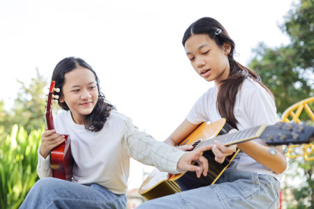 Asian kid girl learning play guitar with friend in the park, Friends playing guitar on picnic in parkの写真素材