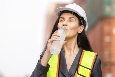 Female Supervisor in Safety Gear Drinking Water at Shipping Yard, Woman in Hard hat Drinking Water at Containers Cargoの写真素材