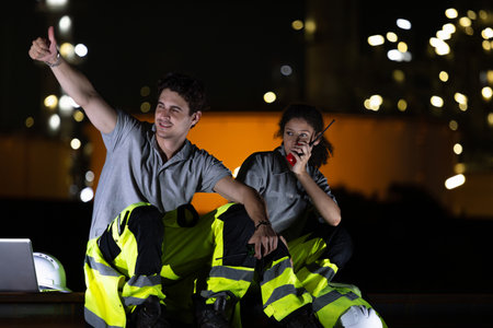 Night Shift Engineers Communicating on Site, Industrial Workers at Night Using Radio and Laptop, Happy Engineers Working Night Shift at Refineryの写真素材