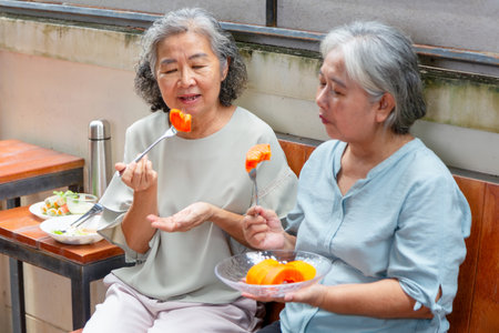 Two Senior Asian Women Enjoying Fresh Papaya Outdoors, Elderly Friends Sharing Healthy Fruit on a Patio, Happy Grandmothers Eating Tropical Fruit Togetherの写真素材