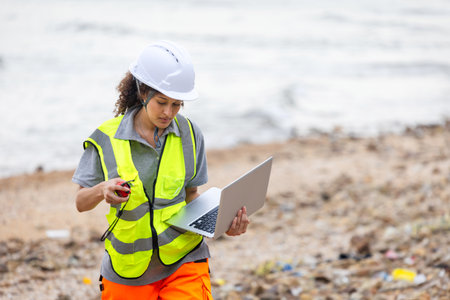 Environmental Scientist Conducting Beach Cleanup Survey, Female Engineer Using Laptop for Coastal Area Assessment, Worker in Safety Vest and Hard Hat Documenting Outdoor Siteの写真素材