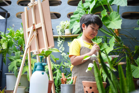Young Asian Boy Painting Outdoors Among Plants, Creative Child with Easel and Brush in a Green Garden, Little Artist Enjoying Painting in a Lush Outdoor Settingの写真素材