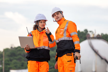 Team of Engineers Conducting Operations at Oil Storage Field Industrial Site, Engineers Collaborating at Industrial Site with Storage Tanks, Industrial Workers Communicating and Using Laptop at Siteの写真素材