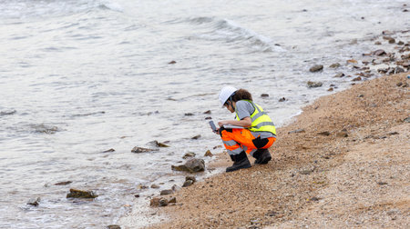 Female Engineer Using Smartphone and Laptop for Coastal Area Assessment, Environmental Scientist Conducting Beach Cleanup Survey, Worker in Safety Vest and Hard Hat Documenting Outdoor Siteの写真素材