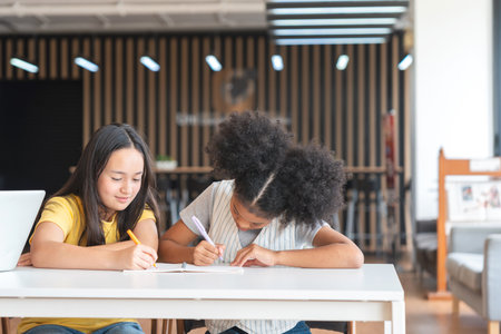 Diverse Young Girls Studying Together at School, Two Elementary Students Focused on Writing in Classroom, Kids Collaborating on Homework at a Deskの写真素材
