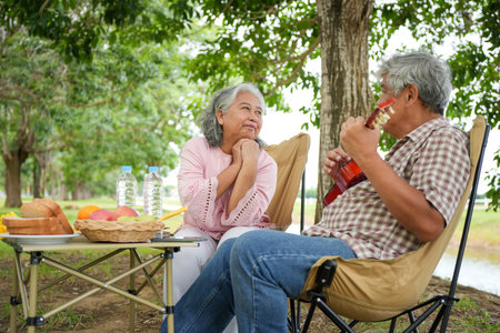 Elderly Asian Couple Relaxing with Ukulele and Picnic in Outdoor Setting, Happy Senior Man Playing Ukulele for Wife in Nature, Joyful Retired Couple Enjoying Music and Togetherness on Outdoor Tripの写真素材