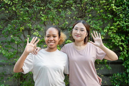 Happy friends waving and smiling outdoors, Two young women posing together and greeting in garden, Cheerful diverse friends standing arm in arm and waving at cameraの写真素材