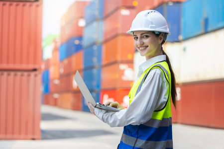 Female engineer using laptop while working at cargo container yard, Smiling industrial worker in safety gear managing logistics at port, Young woman inspecting freight operations with laptopの写真素材