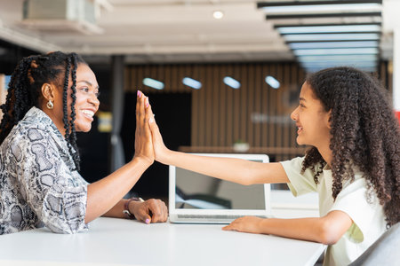 Mother and Daughter High Fiving After Successful Online Learning Session, African American Woman and Girl Celebrating Achievement While Using Laptop, Tutor and Student Giving High Five Over Computerの写真素材