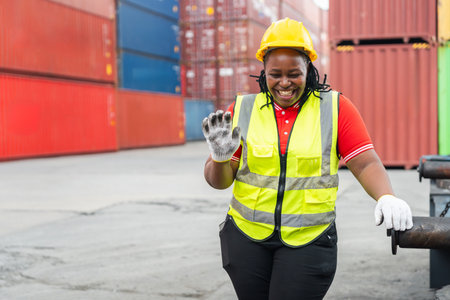 Happy Female Logistics Worker Waving or Signaling Success at Busy Shipping Port, African American Woman in Hard Hat and Safety Vest Smiling Confidently at Workの写真素材