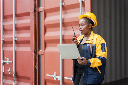 Confident dock worker managing logistics with laptop and radio at shipping terminal, Female industrial worker communicating via walkie talkie while using laptop at container yardの写真素材