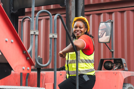 Happy African American Female Worker Climbing Heavy Machinery at Depot, Engineer Woman in Safety Vest and Hardhat Smiling, Portrait of Confident Female Industrial Operator in Cargo Container Yardの写真素材