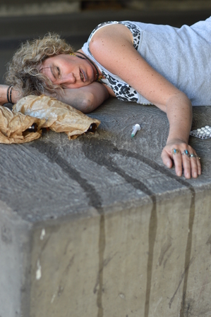 A drunk, homeless woman sleeping under a bridge with bottles of alcohol and other drugs around herの写真素材