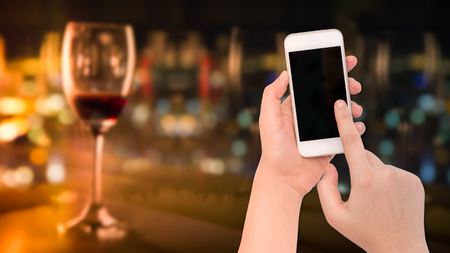 Female  hand holding a phone on blurry wine glass with night business city backgroundの写真素材
