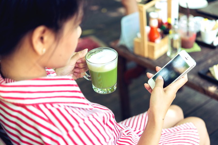 Woman holding Matcha green tea latte and use Mobile Phone on wooden backgroundの写真素材