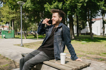 Happy young man wearing blue jeans jacket talking on phone while sitting on a park bench with take...