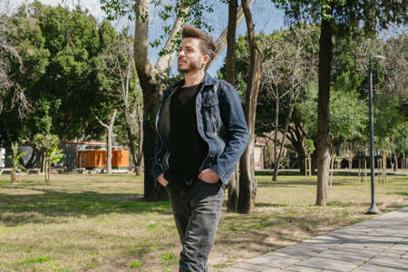 Walking, young man is walking in a green park with his hands in his pockets, a handsome adult wearing a denim jacket, nice weather and springの写真素材