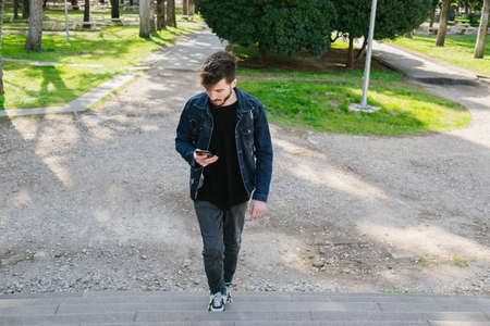 Young man looking at phone at park bench, man in denim jacket looking at black screen, online...