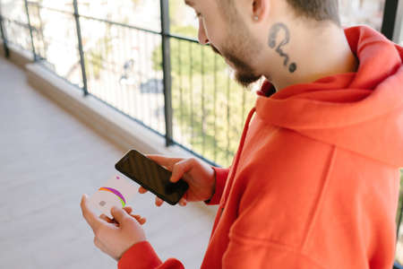 Young man wearing red hoodie holding smartphone with blank white screen and credit card or debit...