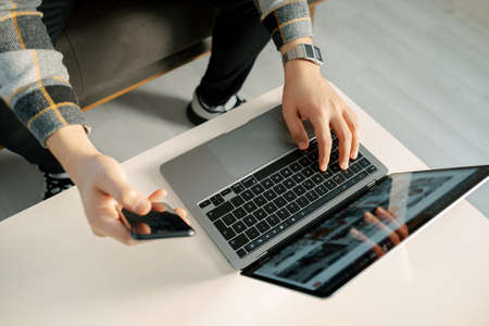 Remote work, young man working on computer standing at table and holding phone in hand, full time...