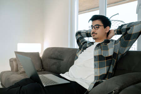 Online meeting, young man displays relaxed demeanor during online meeting with his work team, he is sitting on the sofa with his hands behind his head and the laptop is on his lapの写真素材