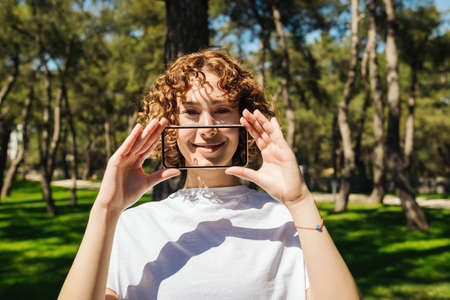 Portrait of a young redhead woman covering her mouth with transparent smartphone screen over green city park background, outdoor. Smiling on phone, digital and social media concepts.の写真素材