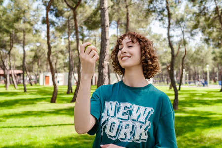 Positive optimistic cheery young redhead woman posing with apple while looking to apple with big smiles at city park. Woman with green apple, against green summer.の写真素材