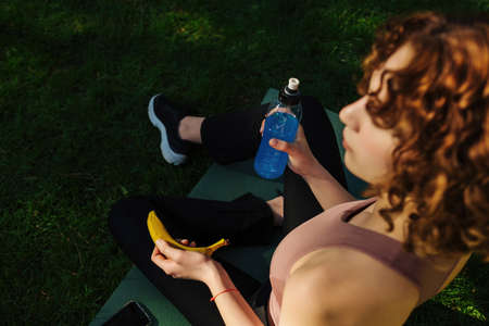 Top view of a woman in fitness wear relaxing after workout sitting on a yoga mat with mobile phone, energy or power drink and banana. Healthy life, organic food and outdoor sport concepts.の写真素材