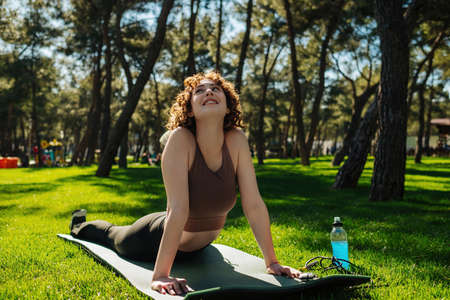 Active young woman in sportswear practicing yoga at city park, outdoors. Laying on fitness mat and...
