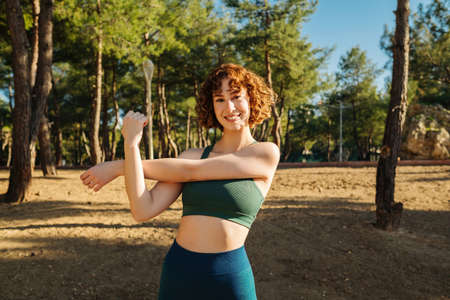 Happy sportive redhead woman wearing sports bra and blue yoga pants standing on city park, outdoors...