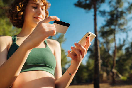 Young redhead woman wearing sportive clothes on city park, outdoors holding mobile phone credit bank...